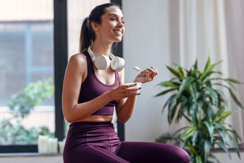 Sporty young woman eating a yogurt while sitting on fitness ball at home.